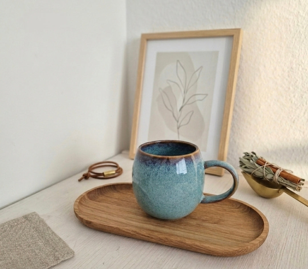 Bauchige Keramiktasse mit blau-türkiser Reaktivglasur und braunem Rand auf einem ovalen Holztablett, dekoriert im modernen Scandi-Stil.